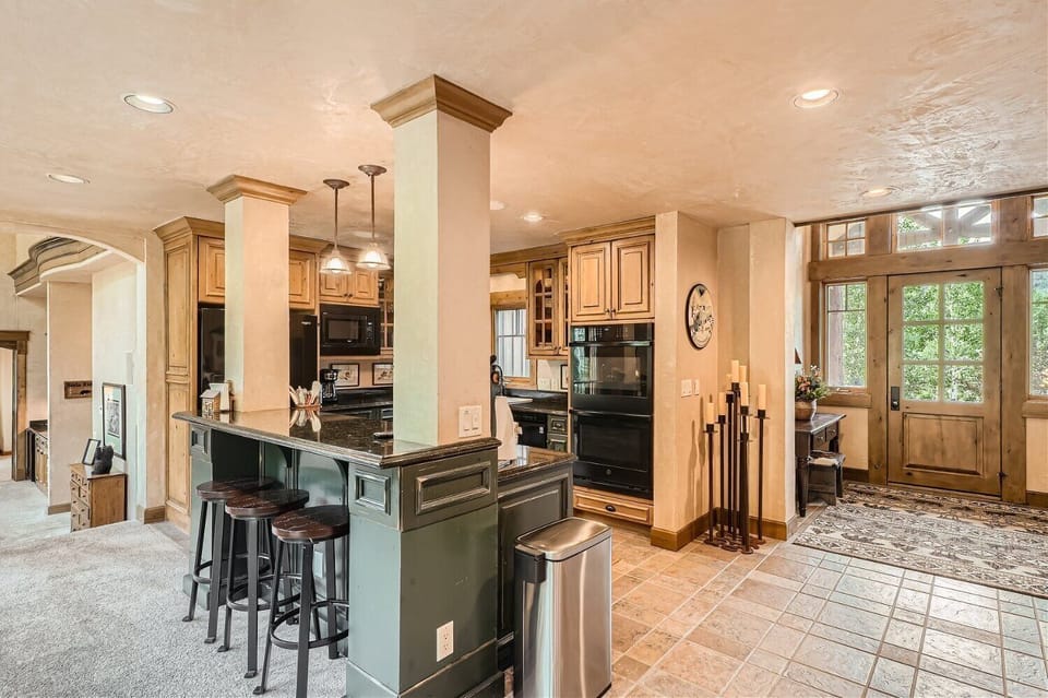 A rustic kitchen with a central island and barstools, wooden cabinets, modern appliances, tiled floor, adjacent to a carpeted entryway with large windows and a wooden front door.