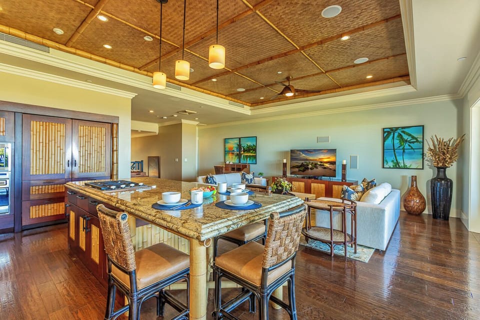 Step into your open-concept living space where island seating flows seamlessly into the lounge. Rich wood floors and a stunning woven bamboo ceiling create warmth, while tropical accents and beach-inspired art set a relaxed island tone throughout.