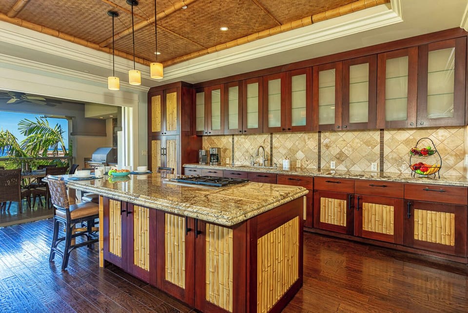 Fully equipped island-style kitchen with rich wood cabinetry, granite countertops, and spacious center island. Opens to outdoor dining and tropical views for seamless island living.