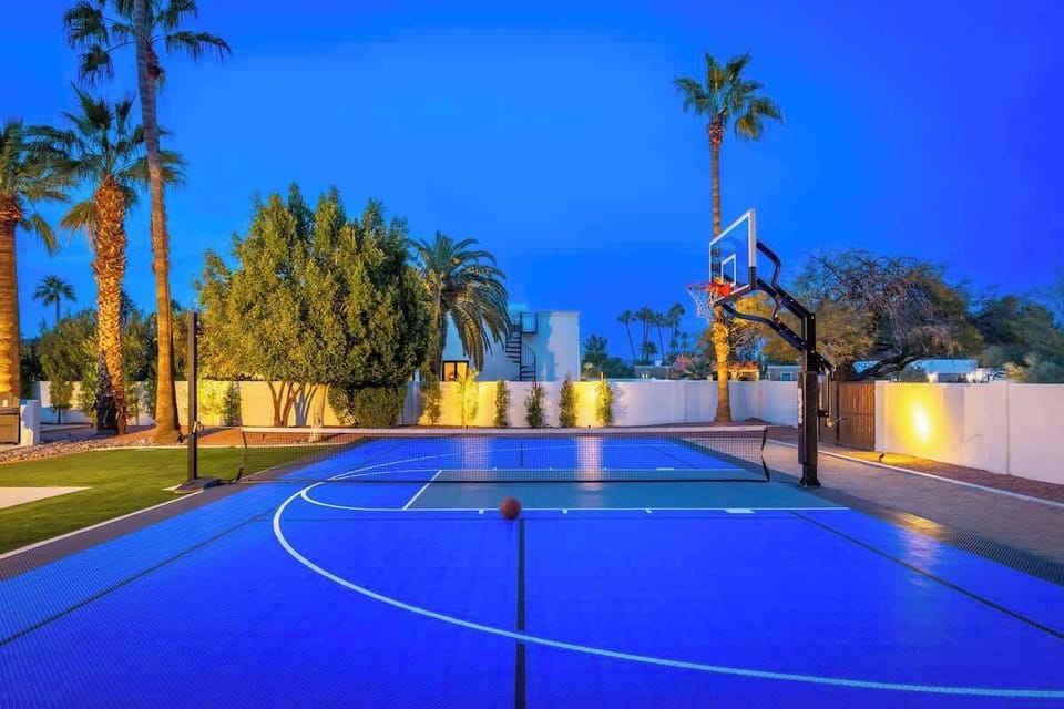 A vibrant outdoor basketball court awaits under palm trees and dramatic twilight skies, perfectly lit for evening games and creating an inviting resort-style atmosphere.