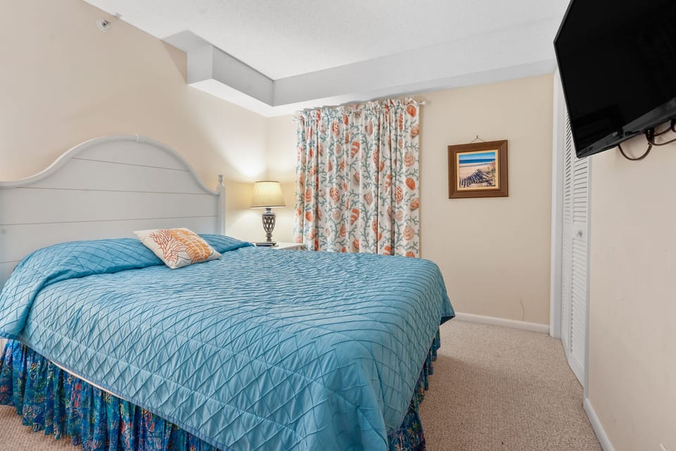 Blue bedding, floral curtains and a mounted TV in a bedroom at Sea Castle 3F, a Crescent Beach vacation rental