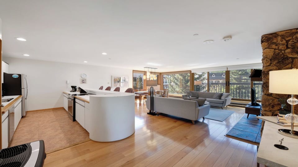 This bright, open-concept space blends kitchen, dining, and living areas, with warm wood floors, a cozy stone fireplace, and windows framing tranquil wooded views.