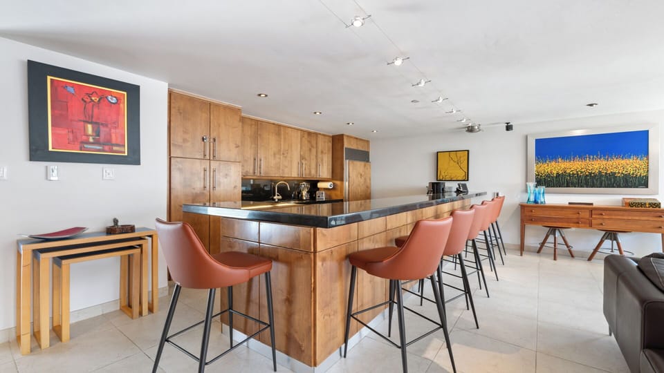 Gather at the sleek breakfast bar with rich wood cabinetry and modern seating, where your morning coffee and conversation flow as easily as the open, art-filled space around you.