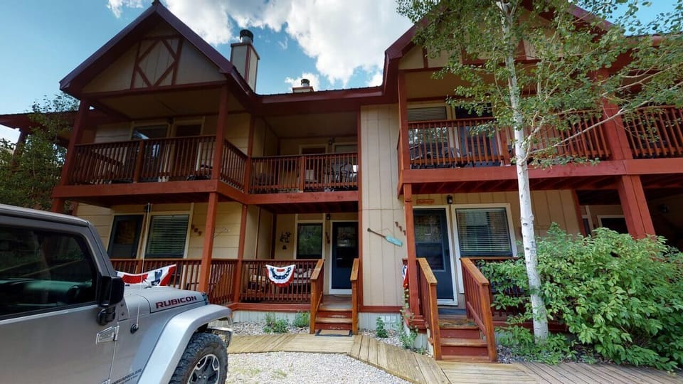 A charming mountain retreat with multi-level decks and warm wood tones, nestled among aspen trees under a bright blue sky. The property features classic alpine architecture with decorative accents and ample outdoor living space.