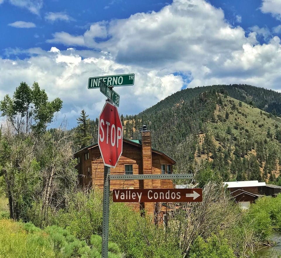 Rustic mountain retreat at the intersection of Inferno Trail and River Road, surrounded by forested peaks and vibrant summer greenery in a quiet alpine valley.