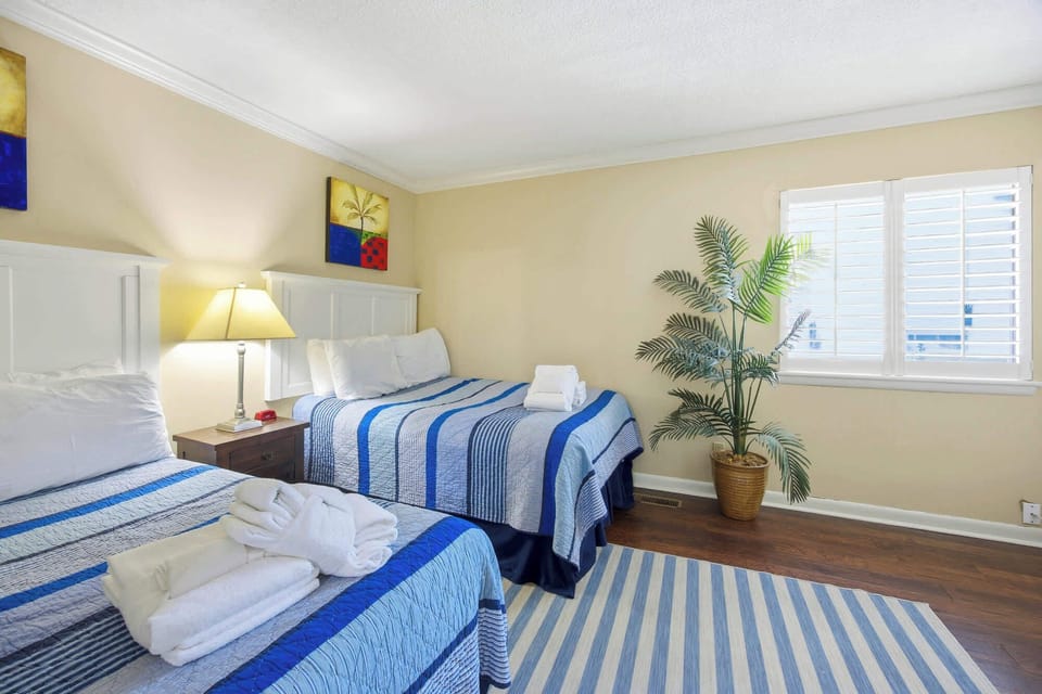 Striped linens in ocean blues pair with a leafy palm to create a cheerful, coastal mood. Bright day beams filter through classic shutters, illuminating the dark wood floors and plush towels waiting for your arrival.