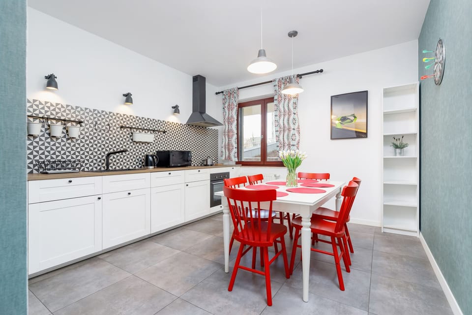 A functional, open-plan kitchen with white cabinets, modern appliances, and a backsplash featuring geometric tiles. The dining area includes a vibrant red dining table with matching chairs, creating a cheerful and inviting space.

