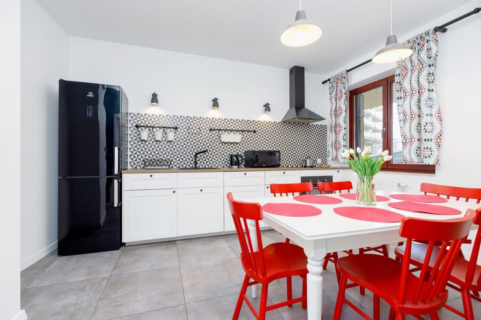 A functional, open-plan kitchen with white cabinets, modern appliances, and a backsplash featuring geometric tiles. The dining area includes a vibrant red dining table with matching chairs, creating a cheerful and inviting space.

