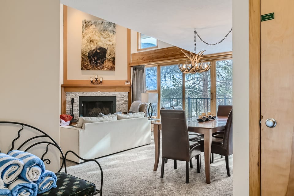 A cozy living and dining area featuring a fireplace, large wall art above it, a white sofa, a wooden dining table with chairs, and large windows with a view of trees. Rolled towels are on a bench.