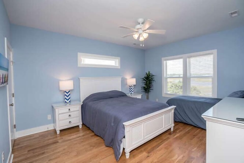 Bedroom with blue walls, two beds, wall-mounted TV, white dresser, ceiling fan, window blinds, bedside lamp, plant, wood floors, and coastal rental style.