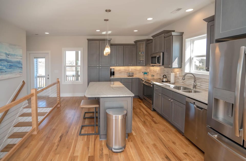 Bright kitchen with gray cabinets, stainless appliances, island seating, pendant lights, wood floors, large windows, and marsh-view style coastal decor.