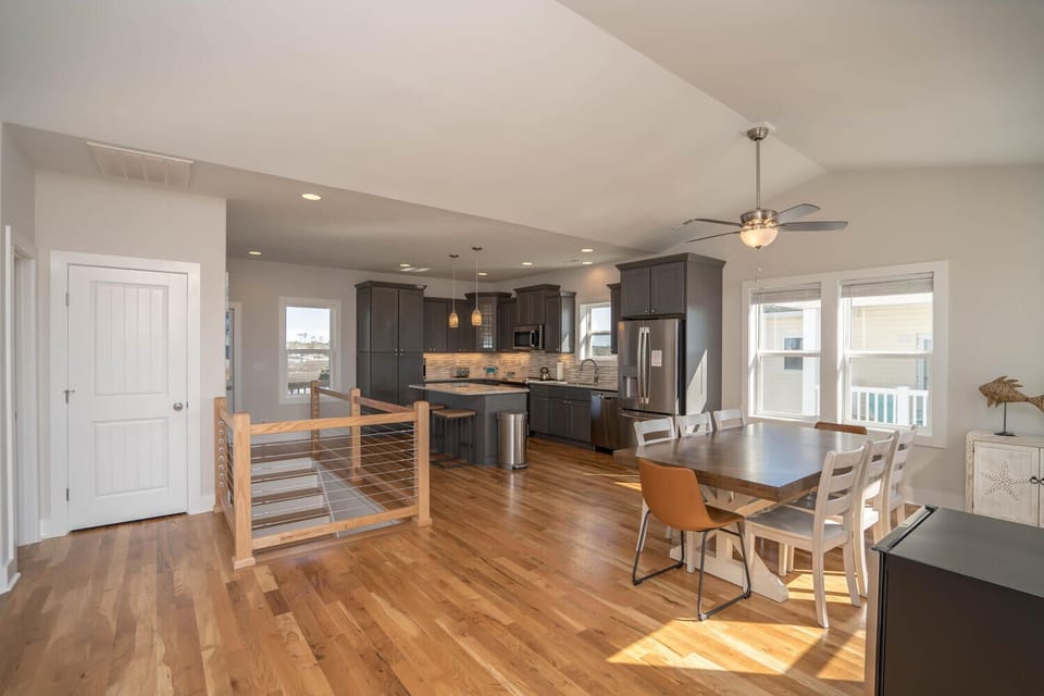 Bright kitchen with gray cabinets, stainless appliances, island seating, pendant lights, wood floors, large windows, and marsh-view style coastal decor.