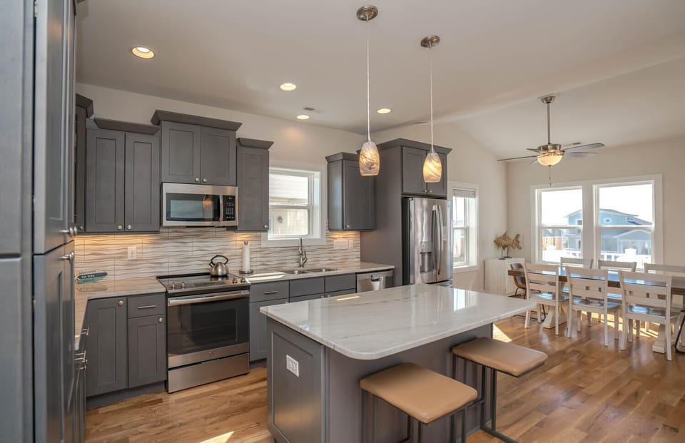 Bright kitchen with gray cabinets, stainless appliances, island seating, pendant lights, wood floors, large windows, and marsh-view style coastal decor.