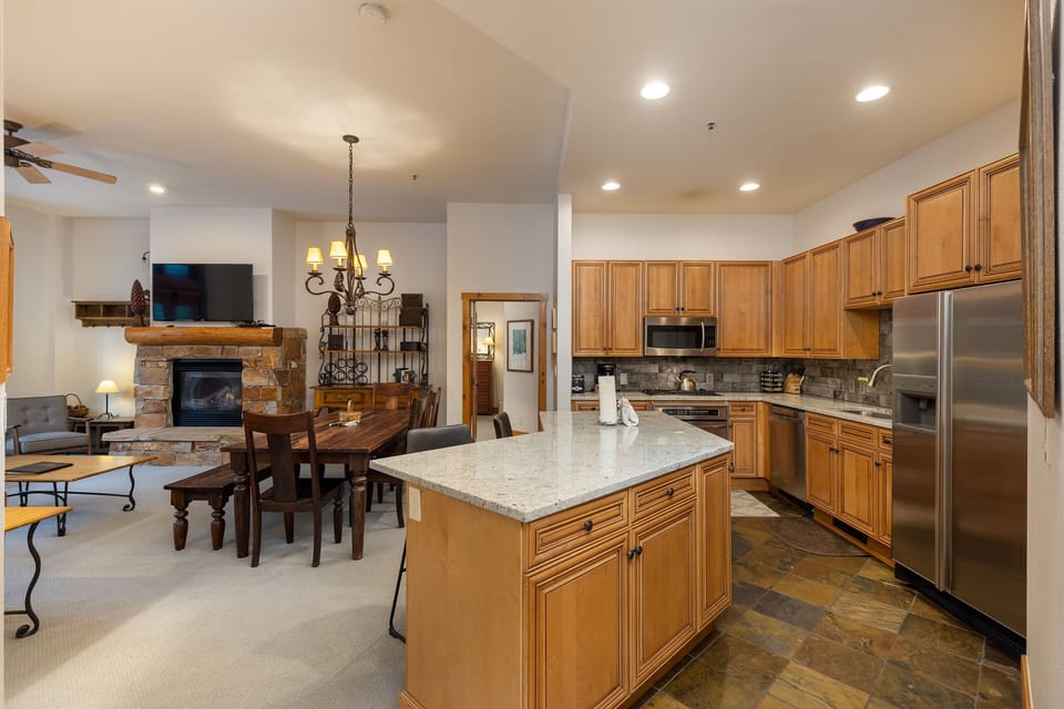 A kitchen and dining area with wooden cabinets, a large island, stainless steel appliances, and a stone fireplace in the corner of the adjacent living room. The space is well-lit and furnished.
