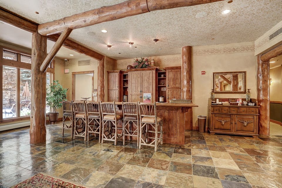 A rustic reception area features a wooden check-in counter with high chairs, a decorative cabinet, and a mirror. The space has stone tile flooring and large windows providing natural light.