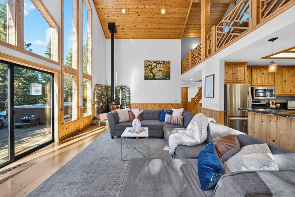 Sunlit living area with vaulted wood ceilings, an oversized sectional, and tall windows framing the pines.