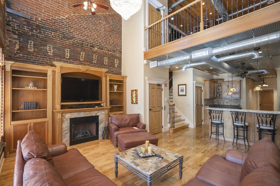 - Nashville loft living room with brick walls and tall ceilings   - Leather seating and fireplace create a warm social space   - Open layout connects to kitchen and upstairs loft area