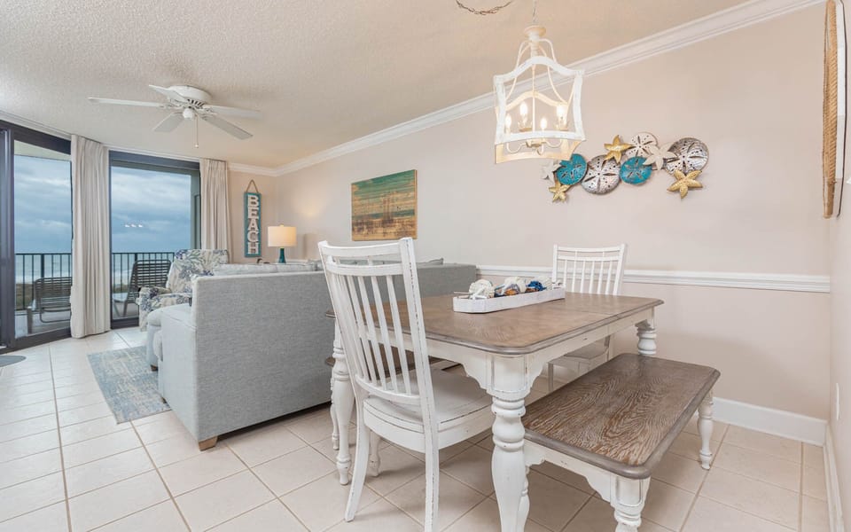 Bright dining area features a wooden table with seating, coastal decor, and large windows offering a view of the beach