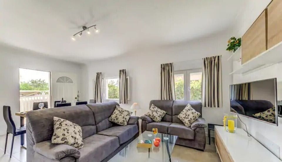 Bright living room with two sofas, glass coffee table, wall-mounted TV and a dining area behind.