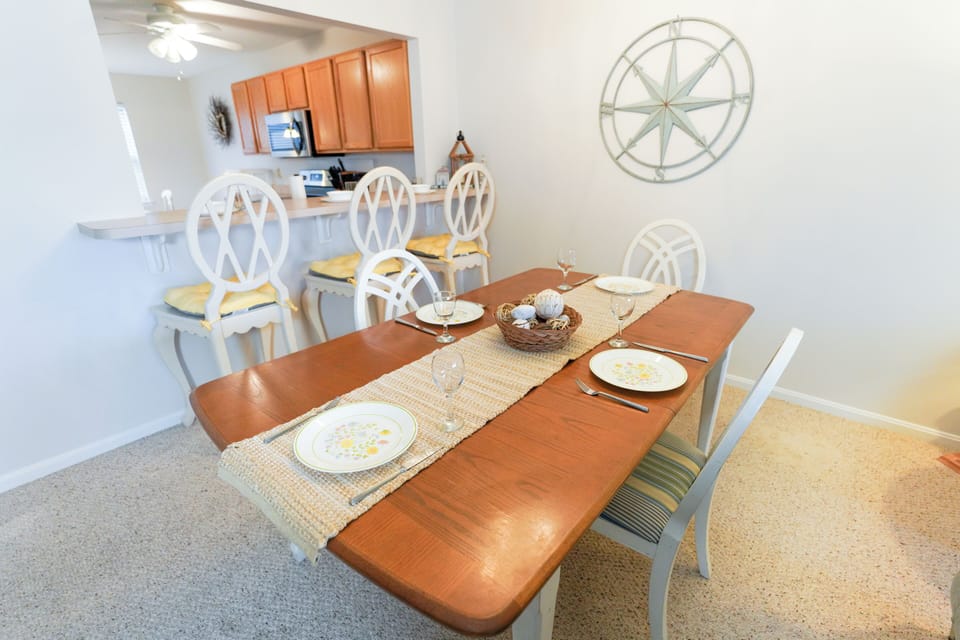 9405 Pettinaro Drive - Dining Area Overview - A well-lit dining space featuring a wooden table set with placemats and plates, situated near the kitchen pass-through and decorated with a wall compass rose.