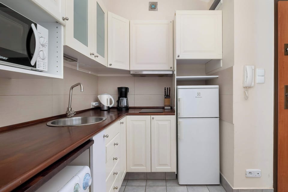 A compact kitchen with white cabinets and brown countertops is arranged in an L-shape. A refrigerator, microwave, and washing machine are integrated into the layout. Light-colored walls make the small space feel bright.