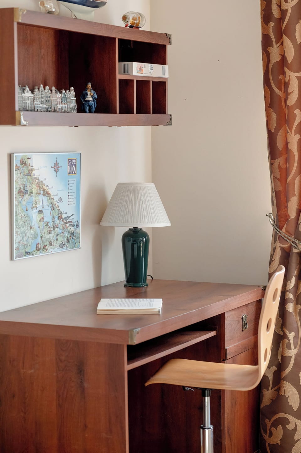 A small work desk with a chair is placed next to shelves in a bedroom corner. A lamp and decorative items sit on the desk. The area serves as a simple study space.