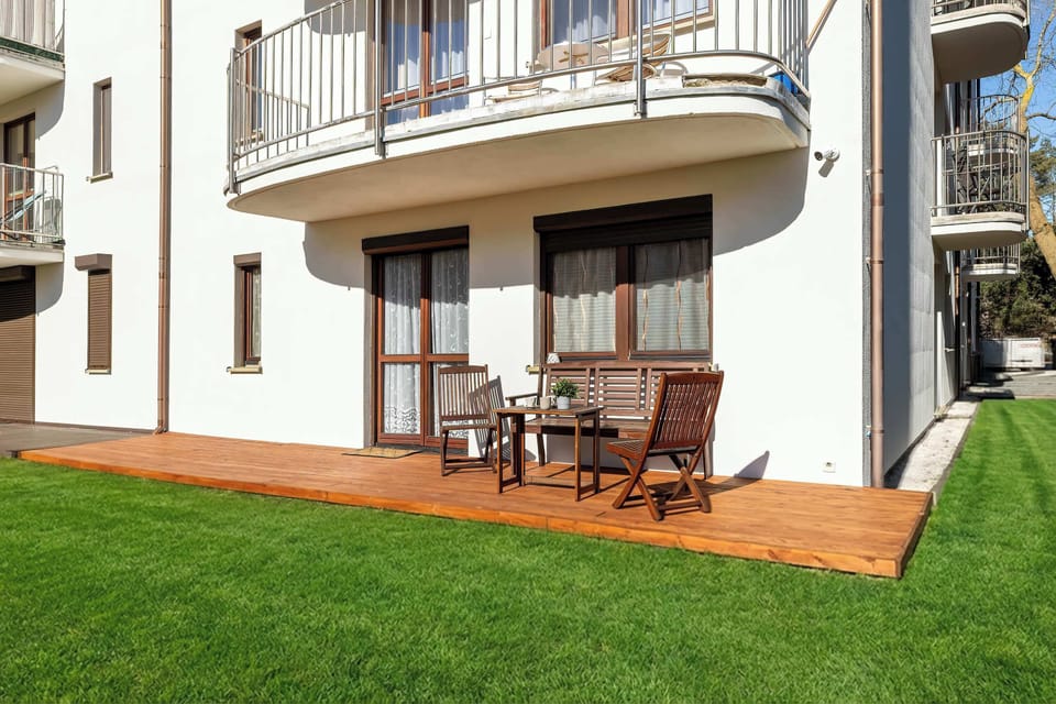 A patio area with wooden decking and outdoor chairs is located outside the apartment. Glass doors lead directly from the interior to the patio. The surrounding lawn adds a pleasant outdoor atmosphere.