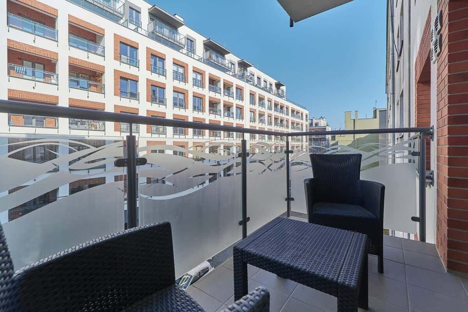A spacious balcony with outdoor seating, offering a picturesque view of neighboring modern buildings and a sunny sky.
