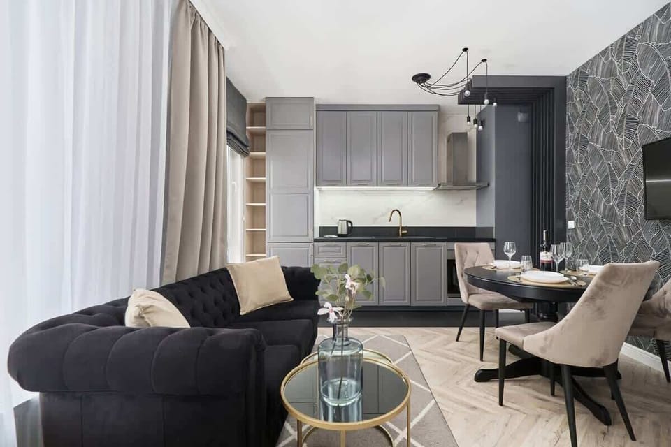 A spacious living room with black sofas, a glass coffee table, and stylish decor. Neutral tones on walls and floors contribute to a chic atmosphere.
