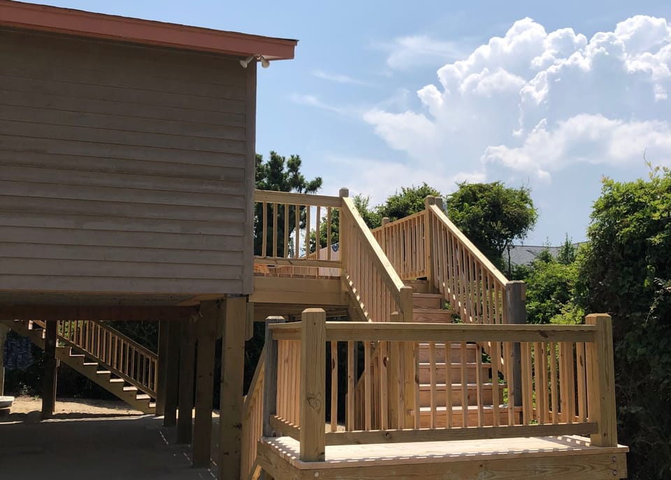 Elevated beach house with wooden deck and stairs surrounded by lush trees beneath a bright sky.