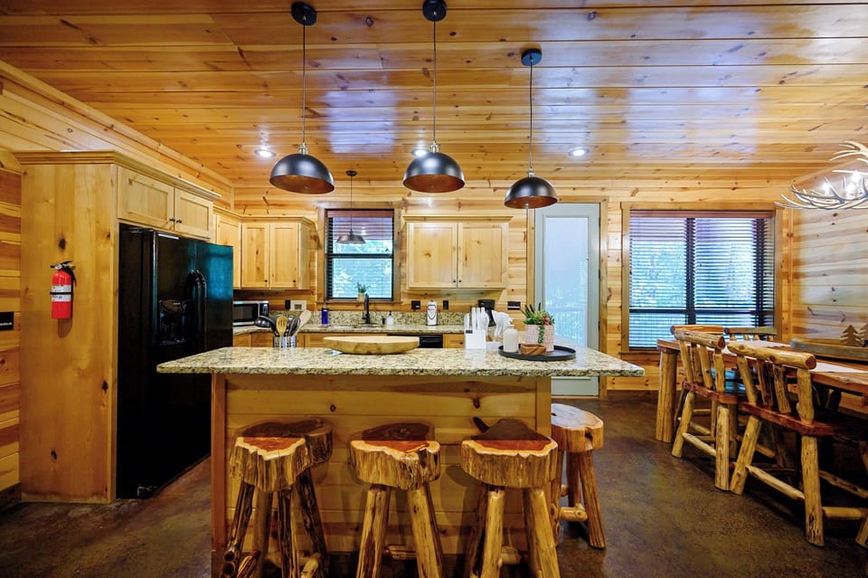 Gather around the rustic wood island with its striking granite countertop, where hand-carved log stools invite you to share meals and memories. The warm pine walls and gleaming pendant lights create a welcoming heart of your mountain retreat.
