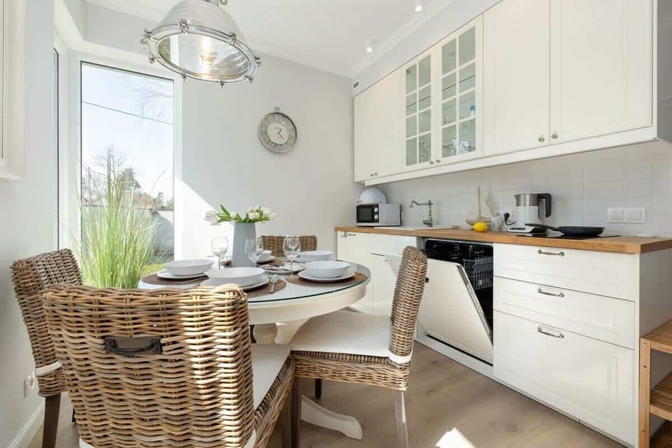 Dining table and chairs in a well-lit living room, styled for a meal. A functional kitchen featuring white cabinets and green plants as decor.