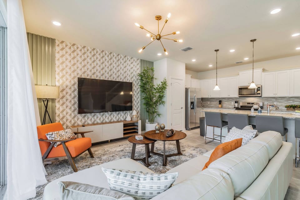 This bright and airy living area combines plush seating and a vibrant orange accent chair for ultimate relaxation, while chic light fixtures and patterned wallpaper bring a contemporary flair to the open layout.
