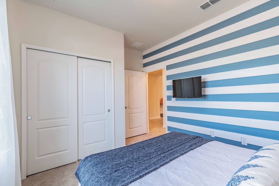 A calming blue-striped accent wall complements the cozy bedding, creating a refreshing vibe in this inviting bedroom. The room features ample natural light and a modern, wall-mounted TV, ensuring a comfortable stay.