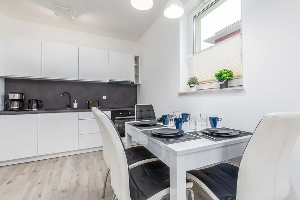 In the foreground is a white dining table with tableware. A kitchenette with white cabinetry, a sleek countertop, and minimalistic decor.