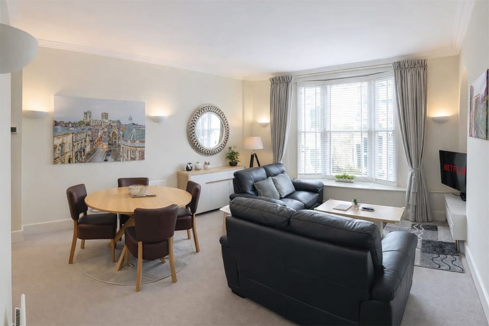 Open-plan living and dining area with black leather sofas, round dining table for four, bay window and York cityscape wall art.