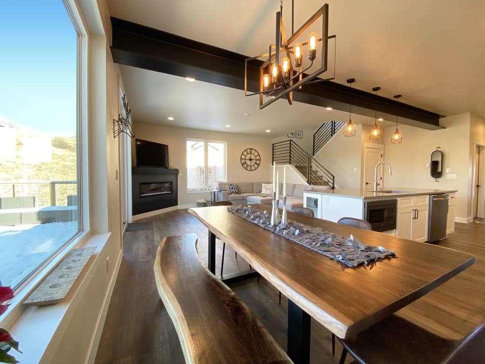 A modern, open-concept kitchen and dining area featuring a wooden dining table, contemporary lighting fixtures, a kitchen island with pendant lights, and a large window offering natural light.