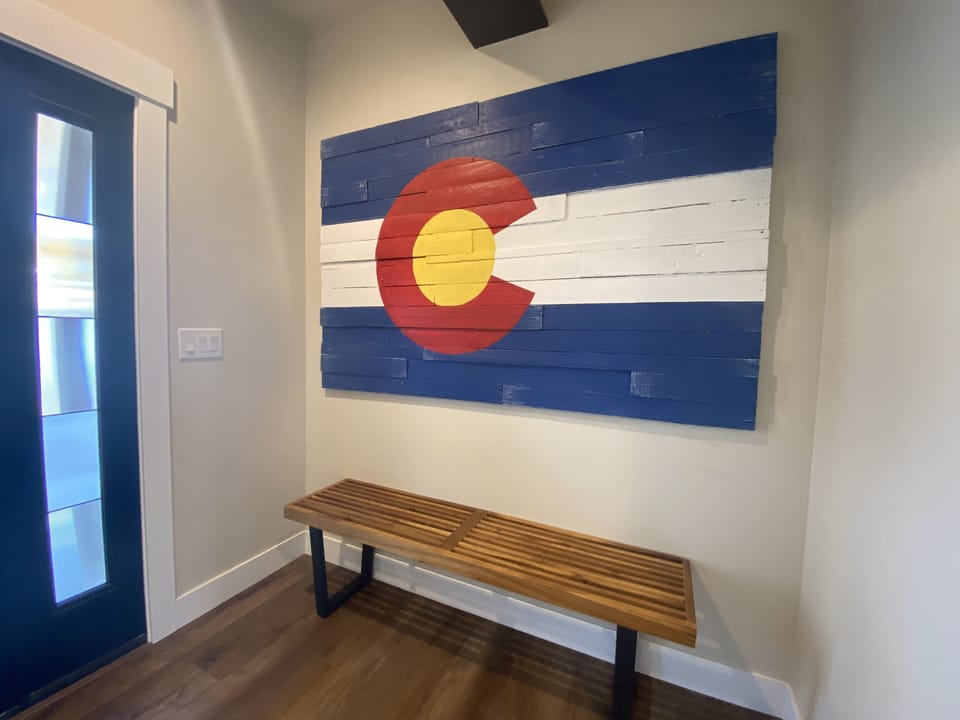 A wooden bench is placed under a large wooden display of the Colorado state flag mounted on a light-colored wall in a room with a dark blue door.