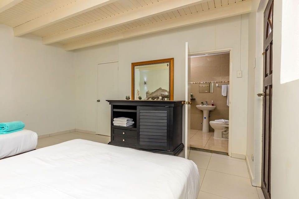 Cozy bedroom with two crisp queen beds, a turquoise towel accent, dark wood dresser, mirror, and a bright en‑suite bathroom framed by white beams and clean tile for a fresh, inviting feel.