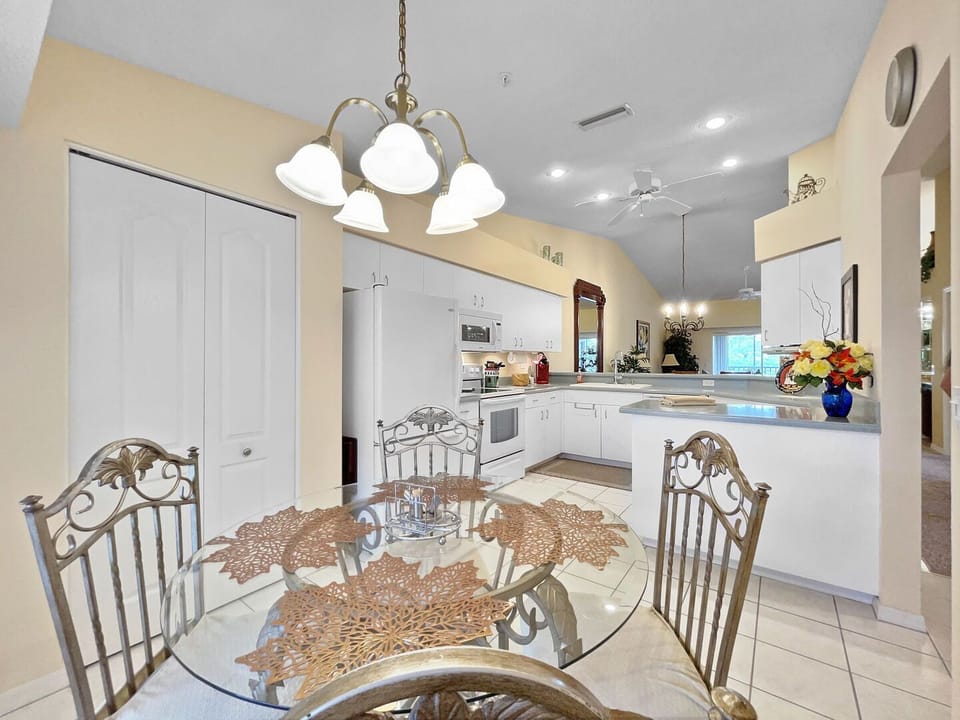 Round glass dining table with iron chairs and open view of the white kitchen with ceiling fan beyond