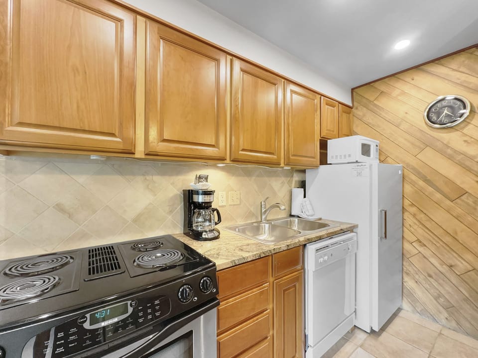 A kitchen with wooden cabinets, an electric stovetop oven, a coffee maker, a sink, a dishwasher, and a white refrigerator. The wall has a clock and wooden paneling.