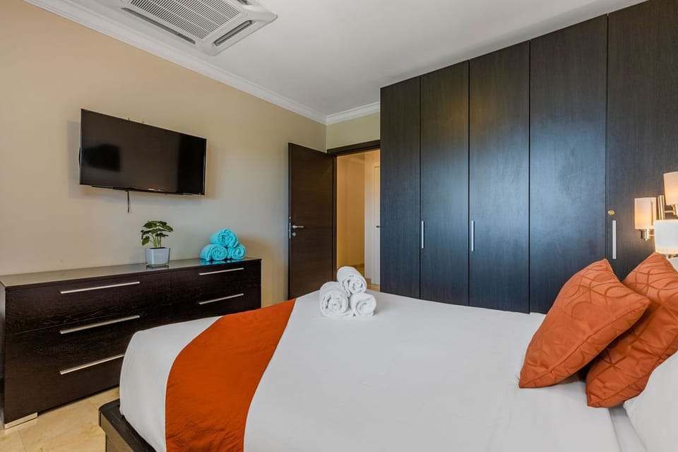 A comfortable bed with air-conditioning unit, a dresser, night stand with lamp TV and closet.