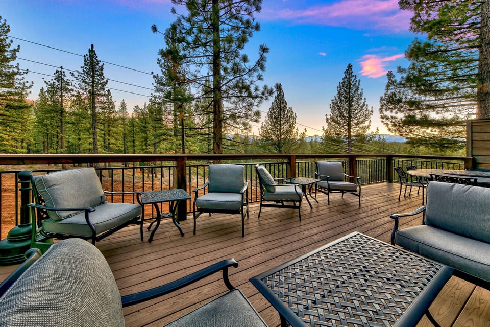 Expansive wooden deck with black metal-framed cushioned chairs and tables, surrounded by pine trees at dusk.