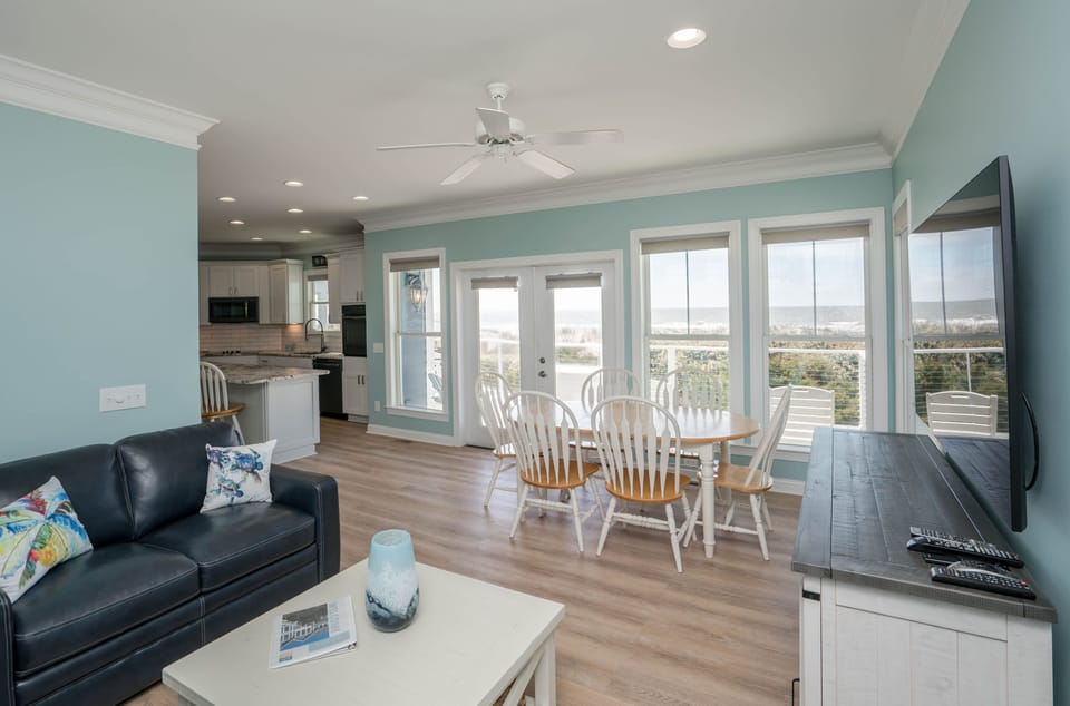 Bright open-concept living and dining area with ocean views, French doors to the balcony, round dining table with seating for six, wall-mounted TV, coastal décor, and a fully equipped kitchen with white cabinetry and granite counters.