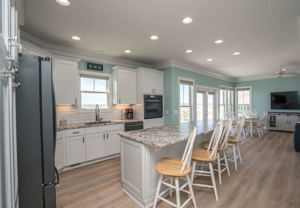 Bright oceanview kitchen with white cabinets, granite island seating for four, stainless appliances, subway tile backsplash, recessed lighting, and open dining and living area overlooking the Atlantic in Oak Island.