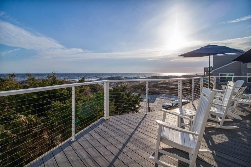 Expansive oceanfront deck with white rocking chairs, cable railings, sun umbrella, and panoramic Atlantic Ocean views over the dunes, perfect for sunrise coffee or sunset relaxation in Oak Island.