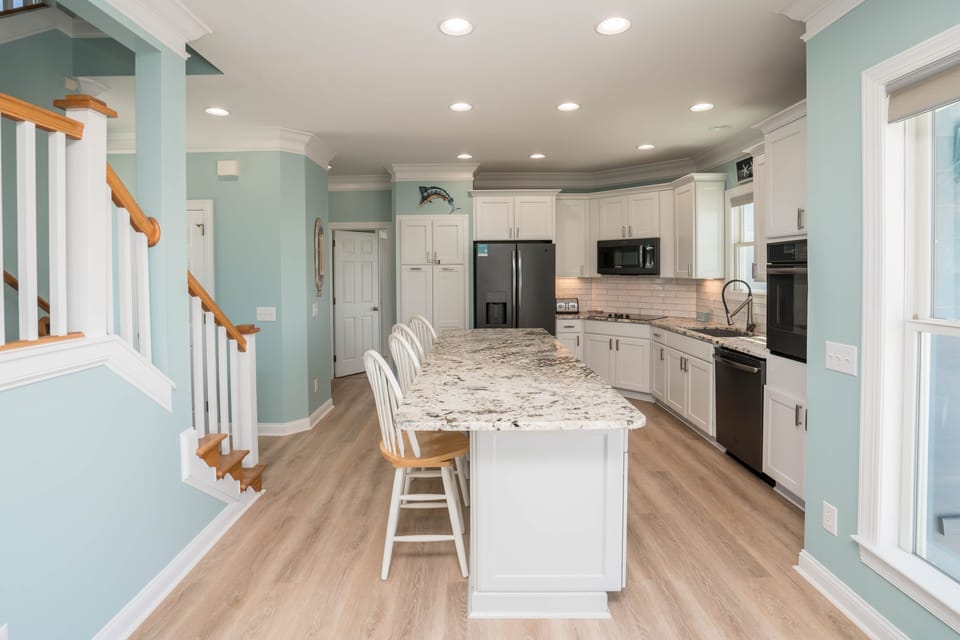 Open-concept coastal kitchen with oversized granite island seating for four, white cabinetry, stainless appliances, subway tile backsplash, recessed lighting, and wood-look flooring in Oak Island.