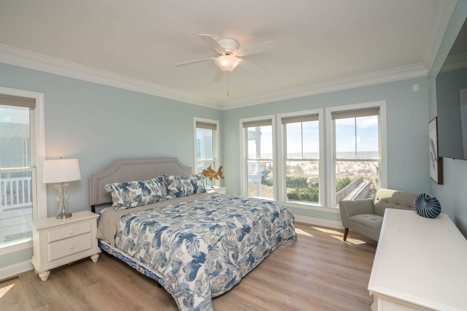 Spacious oceanfront king bedroom with floral bedding, upholstered headboard, nightstands with lamps, ceiling fan, accent chair, dresser with TV, and large windows overlooking dunes and private boardwalk in Oak Island NC.