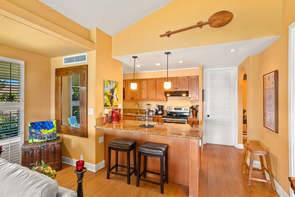 Sunlit open-concept living with a fully equipped kitchen featuring warm wood cabinetry, granite counters, and breakfast bar seating—your perfect space to cook and gather.