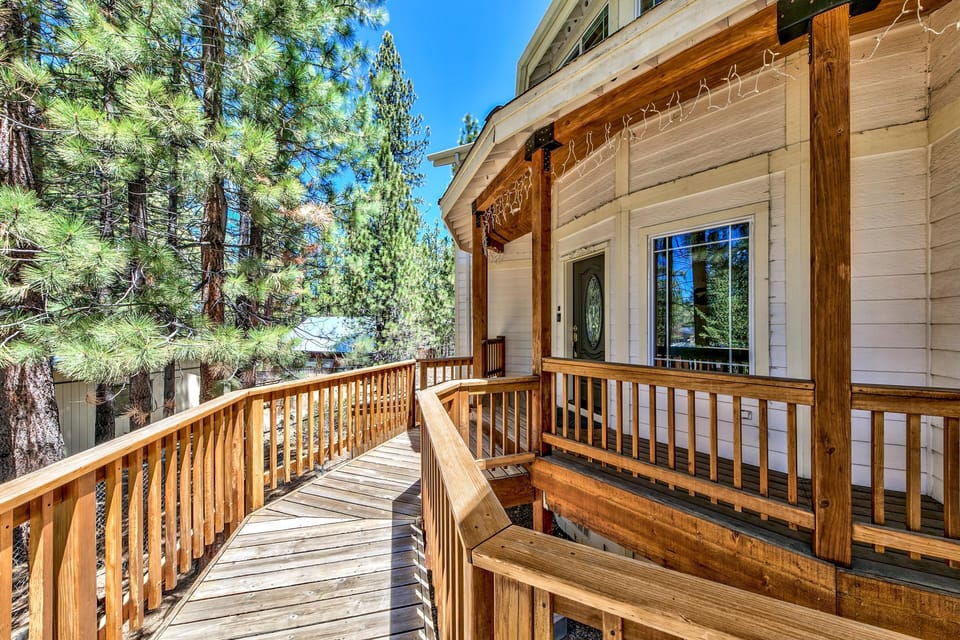 Wide wooden deck wraps around the house entrance, surrounded by tall pine trees under a clear blue sky.